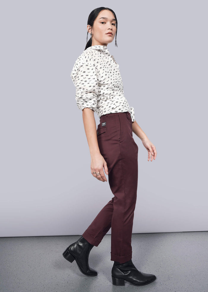 S=A person wearing a white patterned blouse with The Essential Trouser and black ankle boots poses sideways against a plain white background, gazing forward.
