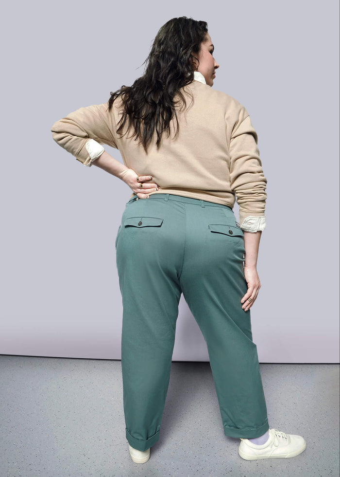 1X= A person with long dark hair, wearing a beige sweater and The Essential Trouser in a relaxed mid-rise fit, stands facing away with one hand on their hip against a light background.