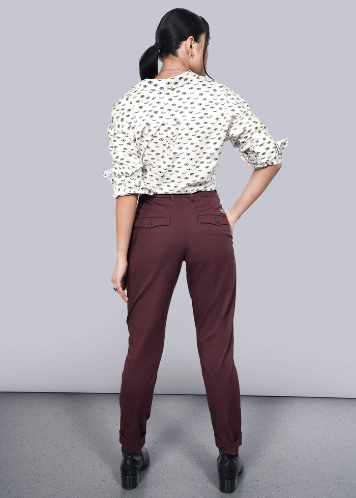 S=A woman stands facing away in The Essential Trouser, maroon with a tapered leg, paired with a white blouse featuring a black pattern. Her hair is tied back, and shes set against a plain white background on a speckled gray floor.