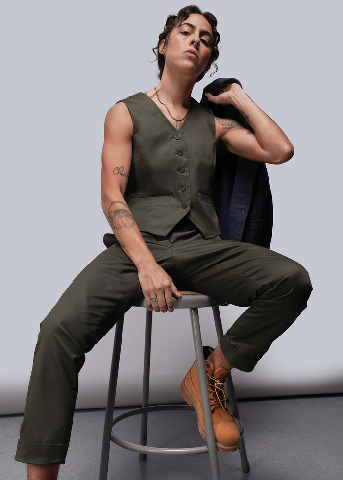 S=Person wearing a green vest and pants sitting on a stool against a plain background