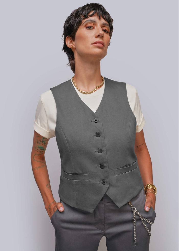 S=A model wearing a dark gray button-up vest over a cream t-shirt, paired with gray trousers and gold jewelry, set against a solid light gray background.