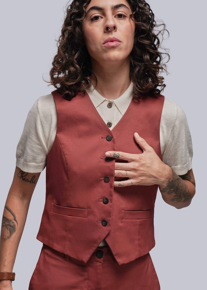 S=Person wearing a rust colored vest over a white sweater polo with tattoos on arms, standing against a plain background