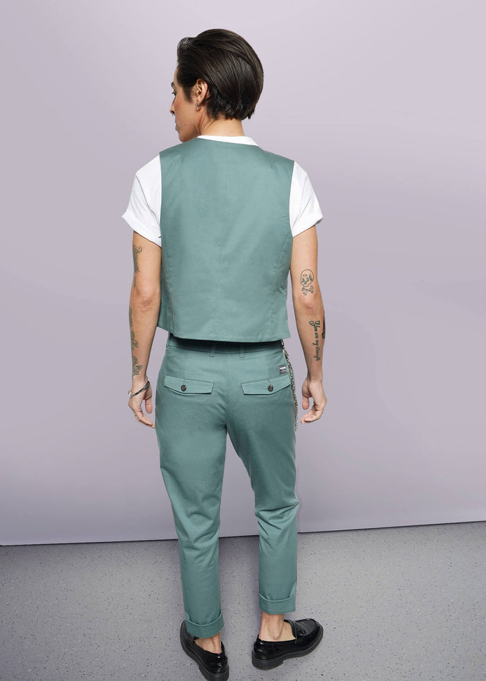 S=A person with short dark hair, wearing The Essential Vest—tailored fit, stretch cotton, 5-button closure—with matching pants, white T-shirt, and black shoes stands facing away from the camera, displaying arm tattoos against a plain white background.