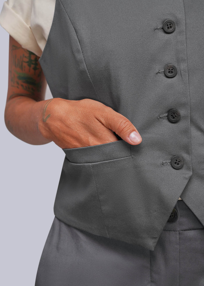 S=Close-up of a person wearing a gray tailored vest with dark buttons and a side pocket, with one hand inserted into the pocket. A cream short-sleeve shirt is partially visible.