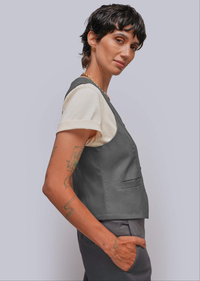 S=A model with short dark hair is shown in a side profile wearing a dark gray sleeveless vest over a cream short-sleeve t-shirt and matching gray trousers, set against a solid light gray background.
