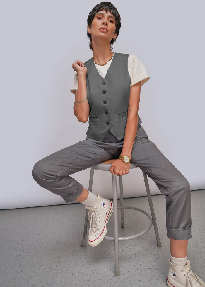 S=A model sits on a metal stool wearing a gray button-front vest, matching gray cropped trousers, a white short-sleeve top, and white high-top sneakers against a light gray studio background.