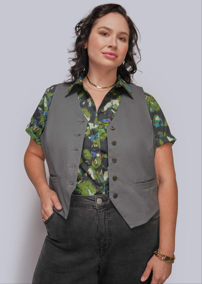 1X=A woman modeling a gray sleeveless vest over a green and blue floral short-sleeve button-up shirt, paired with dark gray jeans, against a light gray studio background.