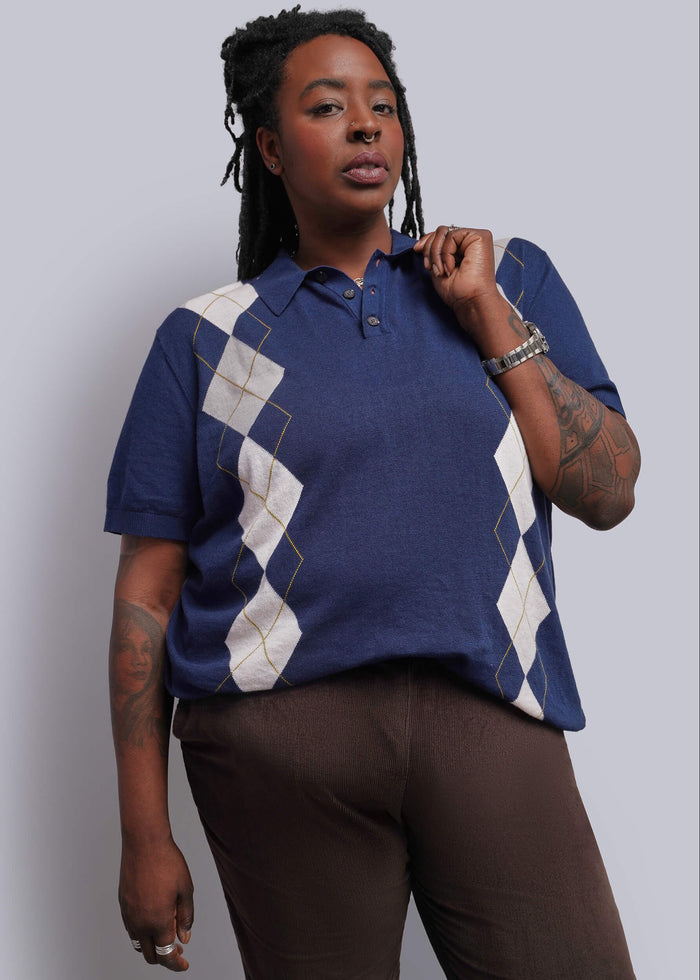 1X=A woman modeling a short-sleeve polo shirt with a navy blue base and white argyle diamond patterns on the chest and sleeves, paired with brown pants, against a solid light gray background.