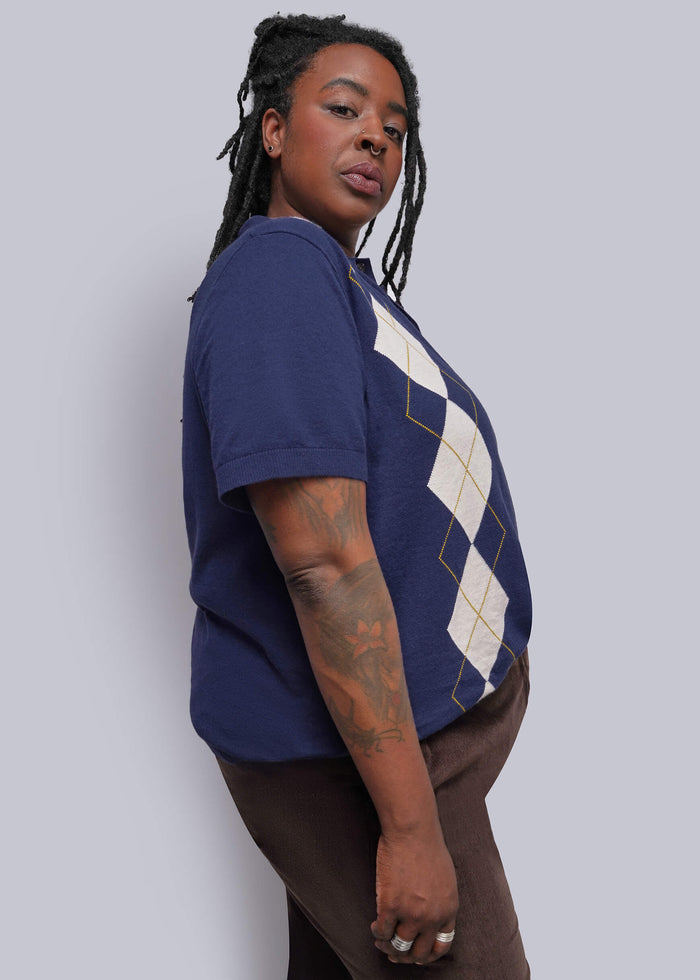 1X=A woman wearing a navy blue short-sleeve t-shirt with a cream and gold argyle pattern on the front, paired with brown pants, standing against a solid light gray background.