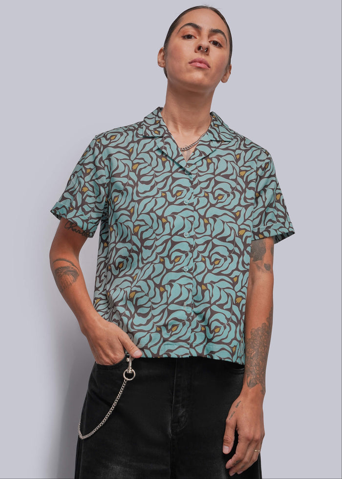 S=A model wearing a short-sleeve button-up shirt with a teal, brown, and mustard yellow abstract pattern, paired with black pants, against a solid gray background.