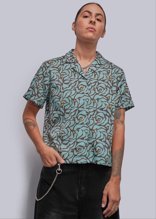 S=A model wearing a short-sleeve button-up shirt with a teal, brown, and mustard yellow abstract pattern, paired with black pants, against a solid gray background.