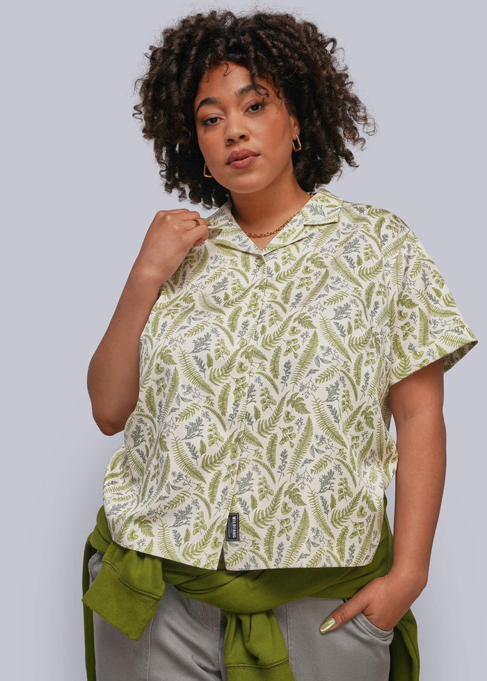 1X=A woman modeling a short-sleeve collared button-up shirt with an olive green and cream fern pattern, styled with gray trousers and a green sweatshirt tied around her waist, against a solid gray studio background.