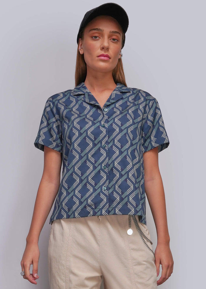 S=A woman modeling a short-sleeve button-up shirt with a blue and beige geometric zigzag pattern, paired with beige pants and a black cap, against a light gray studio background.