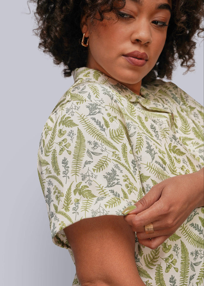 1X=A close-up of a woman wearing a short-sleeve collared shirt with an all-over green fern and leaf botanical print on a cream background, shown against a light gray studio background.