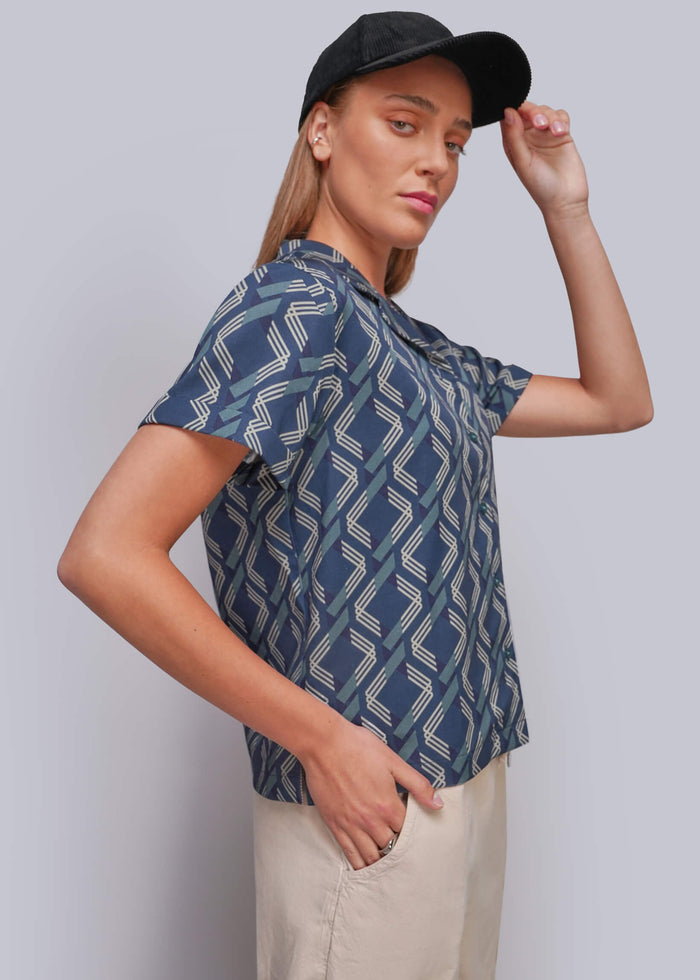 S=A woman modeling a short-sleeve button-up shirt with a blue and beige geometric zigzag pattern, paired with beige trousers and a black baseball cap, against a solid gray studio background.
