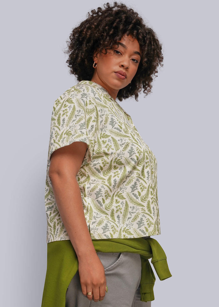 1X=A woman modeling a short-sleeve top with a green and cream botanical pattern, styled with a matching green shirt tied around her waist and gray trousers, against a solid light gray background.