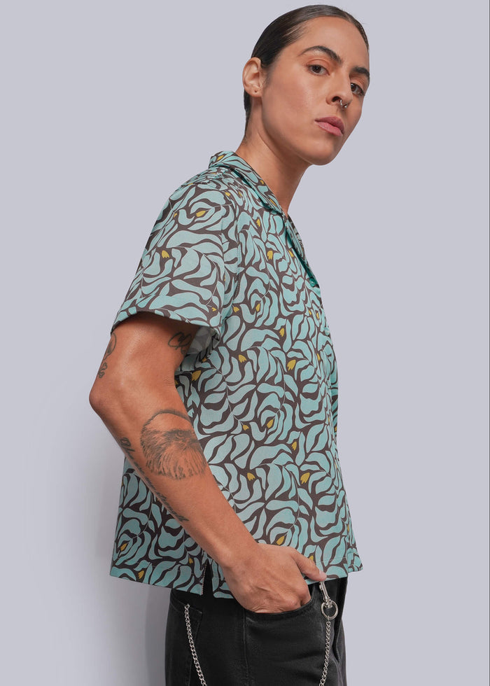S=A model wearing a short-sleeve button-up shirt with a green, brown, and yellow abstract organic pattern, paired with black pants and a silver wallet chain, set against a solid light gray background.