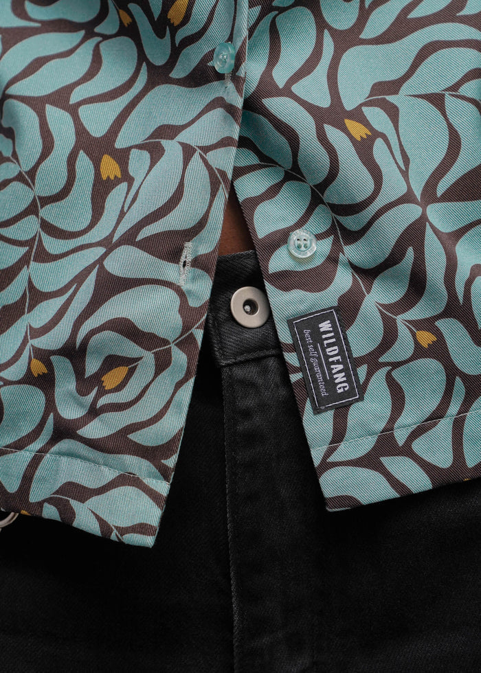 S=Close-up of a button-front shirt with an abstract wavy pattern in light aqua and dark brown, with small mustard-yellow accents. A black fabric brand tab reading WILDFANG is visible near the placket, and the shirt is styled over a black garment.