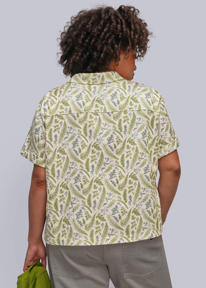 1X=A person shown from the back wearing a short-sleeve button-up shirt with an olive green and cream leafy pattern, paired with gray trousers, against a solid light gray background.