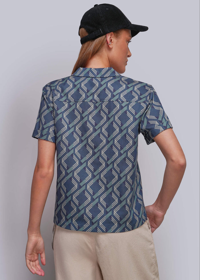 S=A man wearing a short-sleeve button-up shirt with a blue, gray, and white geometric pattern, paired with beige pants and a black baseball cap, shown from a back view against a light gray studio background.