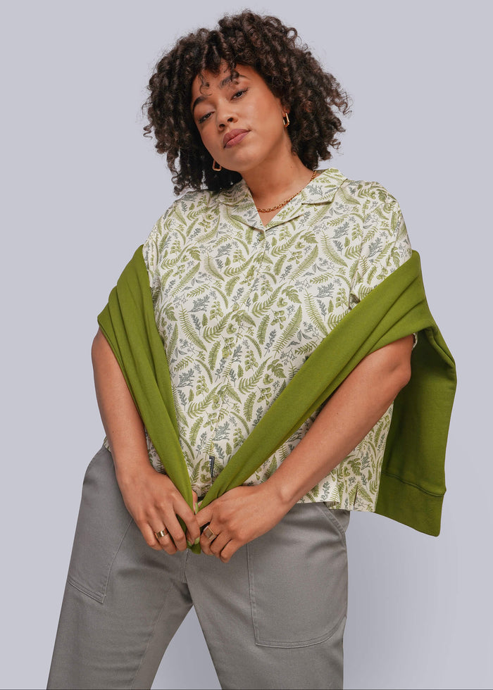 1X=A woman modeling a short-sleeve collared blouse with a green and beige botanical pattern, styled with a solid olive green sweatshirt draped over her shoulders and gray trousers, against a solid gray studio background.