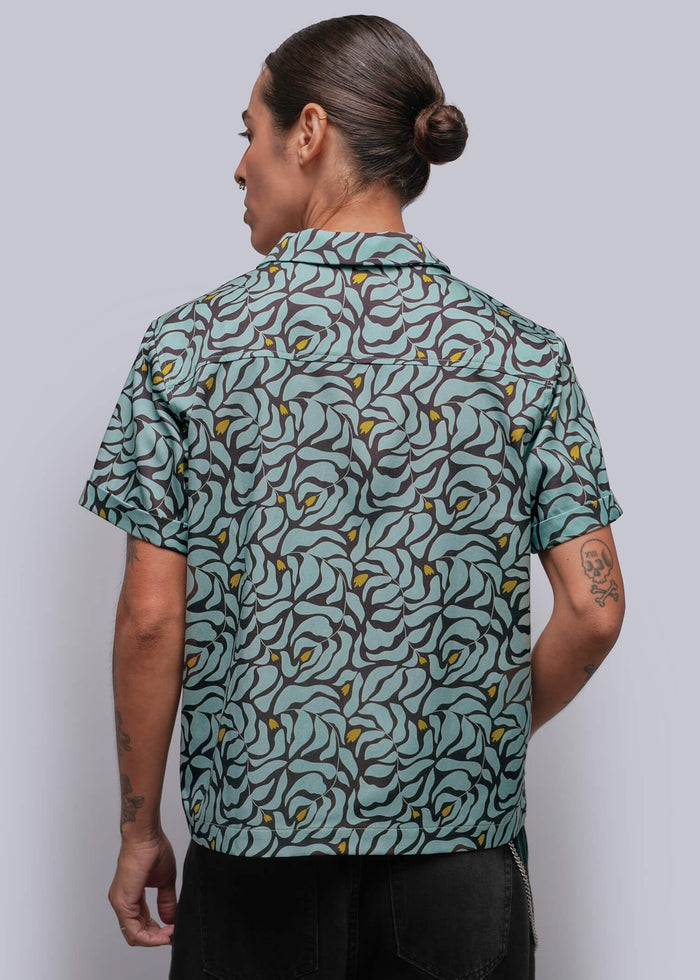 S=A person viewed from the back wearing a short-sleeve button-up shirt with a teal, gray, and yellow abstract floral pattern, paired with black pants, against a solid gray studio background.