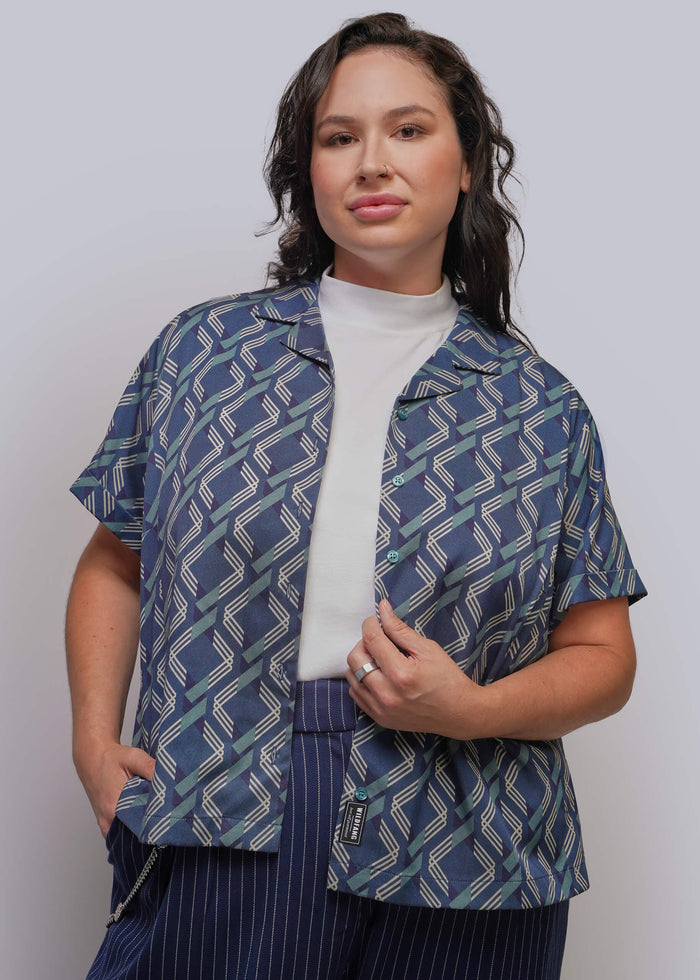 1X=A woman modeling a short-sleeve button-up shirt with a blue, white, and green geometric pattern, worn over a white mock neck top and paired with navy pinstriped trousers, against a light gray studio background.