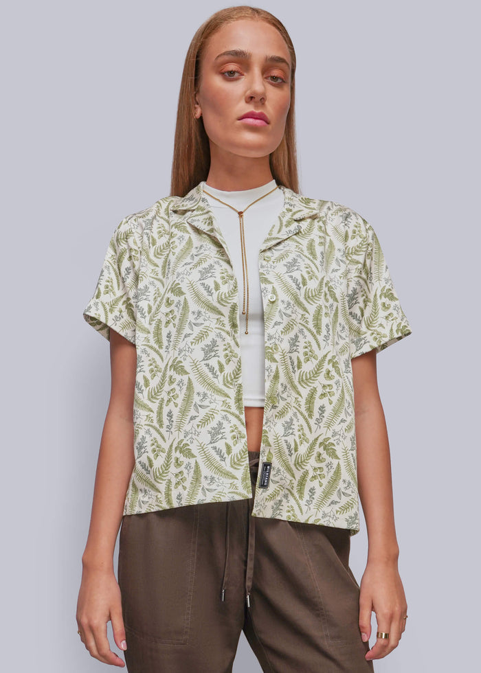 S=A woman modeling a short-sleeve button up with a green and white pattern, worn unbuttoned over a white crop top and brown pants, against a solid gray background.