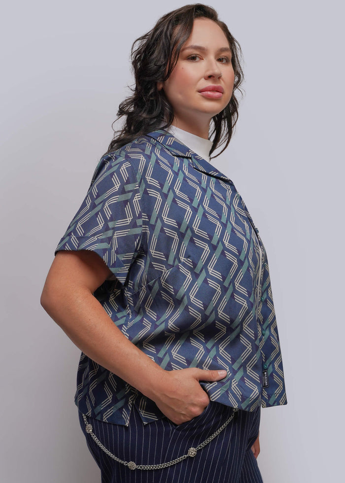 1X=A woman modeling a short-sleeve button-up shirt with a blue, gray, and white geometric pattern, paired with navy pinstriped trousers and a silver chain wallet chain, against a light gray studio background.