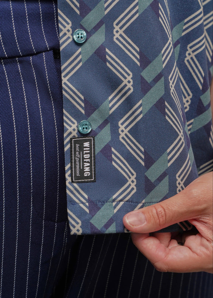 1X=Close-up of a button-down shirt with a blue, teal, and cream geometric pattern, held open by a hand to show the inside collar and a black Wildfang brand label.