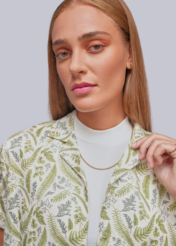 S=A close-up of a woman wearing a white ribbed mock neck top under a short-sleeve button-up shirt with an all-over green fern and leaf botanical print, set against a solid light gray background.