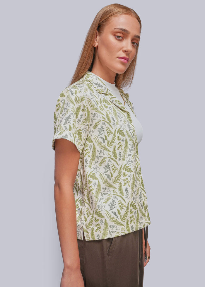 S=A woman wearing a short-sleeve button-up shirt with an olive green and cream botantical pattern, paired with brown trousers, shown against a solid light gray background.
