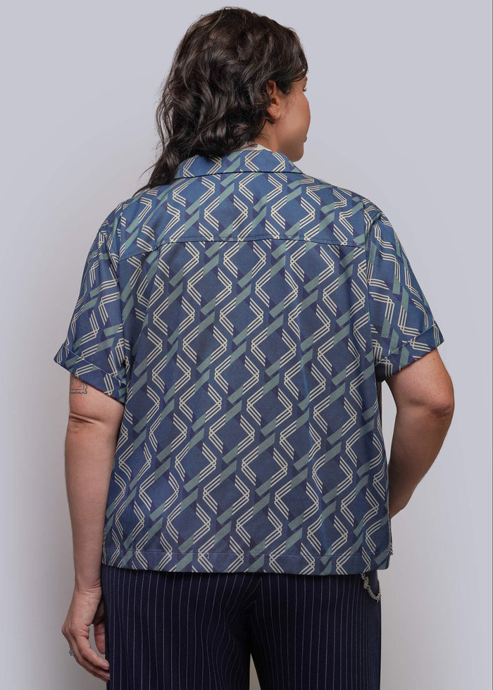 1X=A person shown from the back wearing a short-sleeve button-up shirt with a blue, gray, and white geometric pattern, paired with dark pinstriped pants, against a plain light gray studio background.