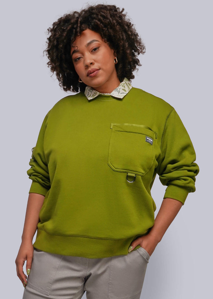 1X=A woman modeling a green crewneck sweatshirt with a decorative cream patterned collar, a chest pocket with a small metal snap detail, and light gray pants, set against a solid gray studio background.