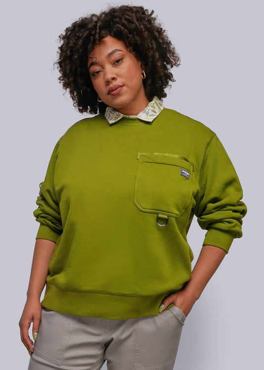 1X=A woman modeling a green crewneck sweatshirt with a decorative cream patterned collar, a chest pocket with a small metal snap detail, and light gray pants, set against a solid gray studio background.