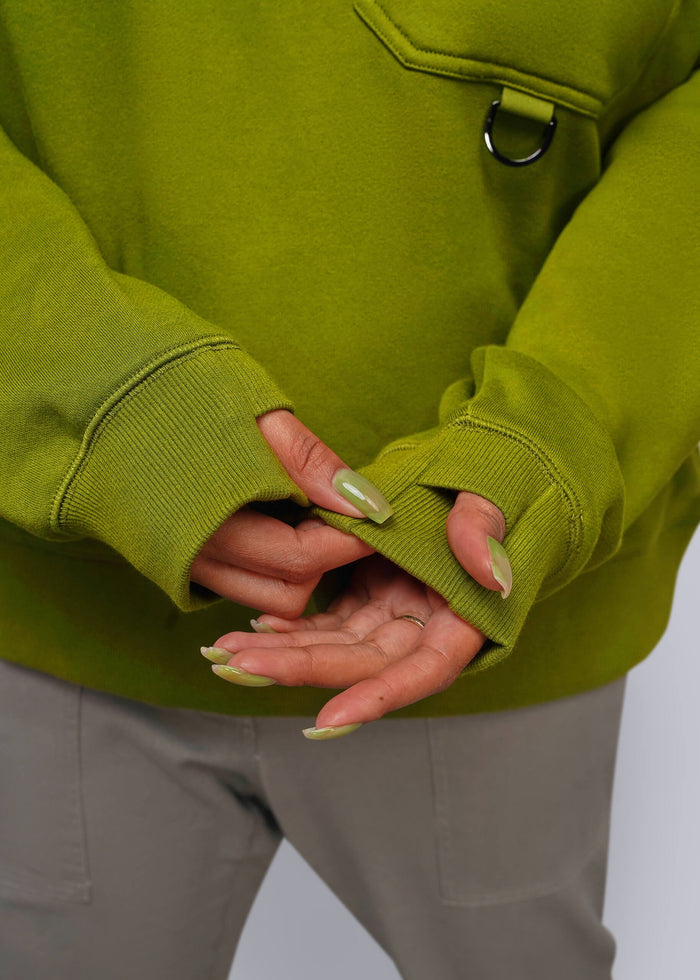 1X=Close-up of a person wearing a green sweatshirt with ribbed cuffs and a small black metal ring detail on the chest pocket, paired with light gray pants.