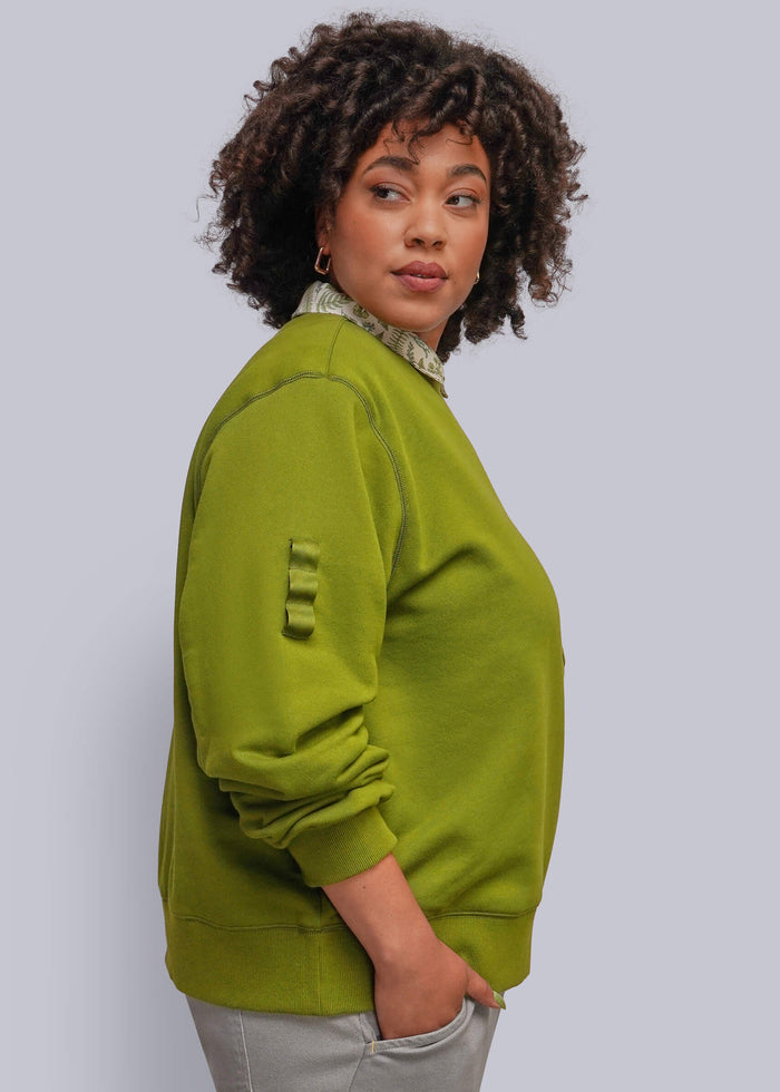 1X=A woman with curly dark hair is shown in profile wearing an olive green crewneck sweatshirt with a unique ribbed sleeve detail and light gray pants, set against a solid light gray background.
