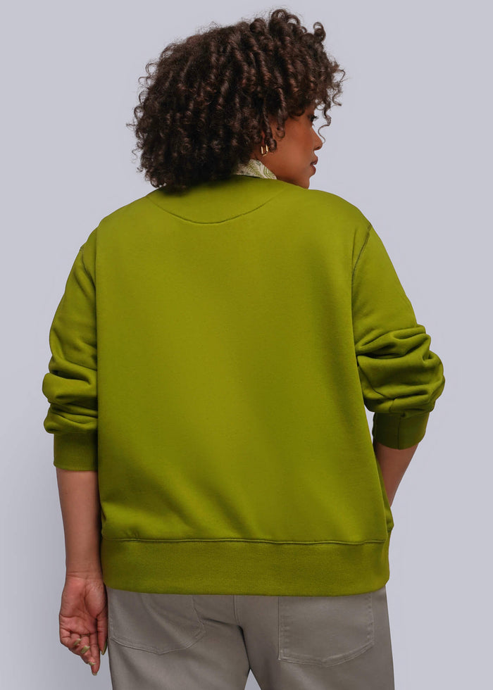 1X=A person shown from the back wearing an green crewneck sweatshirt and light gray pants, standing against a solid light gray studio background.