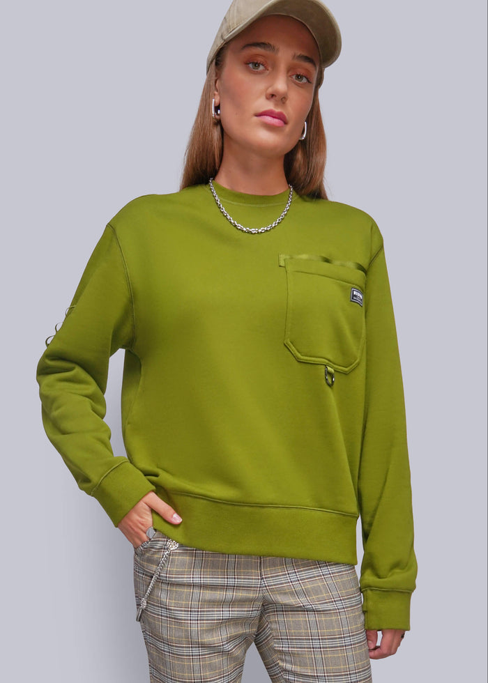 S=A woman wearing a green crewneck sweatshirt with a chest pocket, paired with beige plaid pants and a beige cap, against a solid gray studio background.