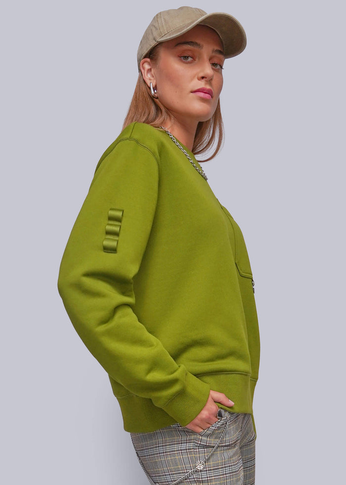S=A woman wearing a green crewneck sweatshirt with unique ribbed sleeve detailing, paired with plaid pants and a beige cap, shown against a solid gray background.