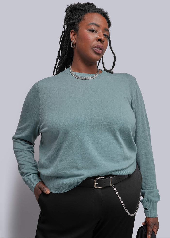 1X=A woman modeling a teal long-sleeve knit top with black trousers, a silver chain belt, and silver jewelry against a solid light gray background.