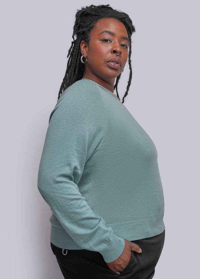 1X=A woman with braided hair is shown in profile wearing a muted teal long-sleeve top and black pants against a plain light gray studio background.