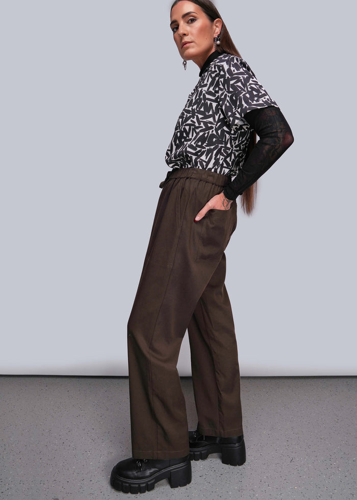 S=Woman wearing a patterned shirt and brown pants against a plain background