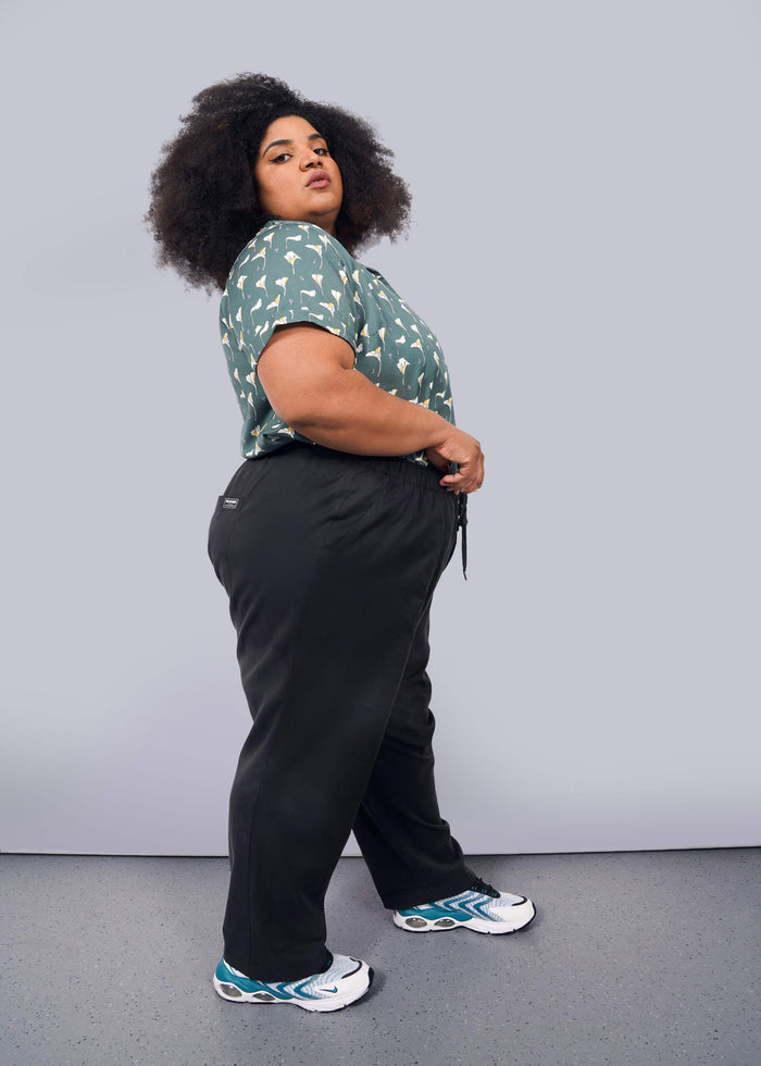 1X= A person with curly hair stands sideways against a white background, wearing a green patterned shirt, The Everyday Elastic Waist Pant in black, and white sneakers with blue accents.