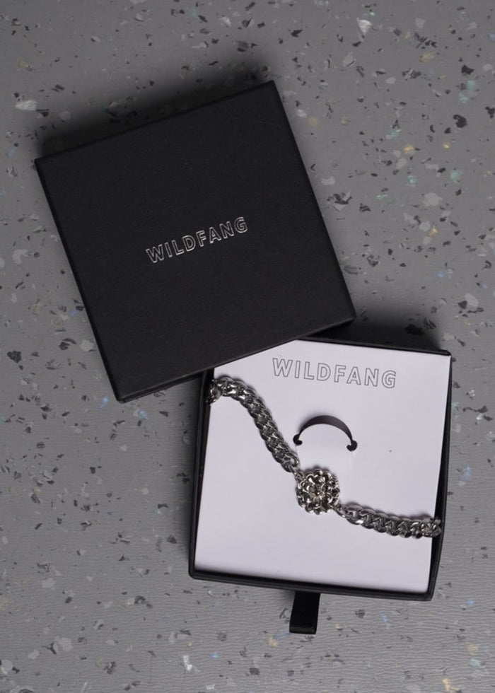 A silver-toned wallet chain with a floral charm, displayed in an open box with the brand name WILDFANG, next to the box lid on a speckled gray surface.