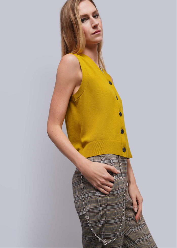 A woman wearing a sleeveless mustard yellow knit vest with dark buttons and plaid trousers with a silver metal wallet chain, posing against a light gray studio background.
