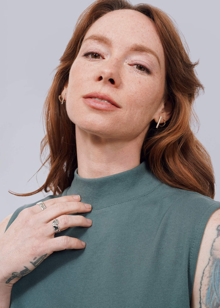 S= A woman with wavy red hair and visible hand tattoos confidently gazes at the camera, wearing The Essential Mock Neck Tank in green. She has light freckles and subtle makeup, with her hand resting gently on her chest.