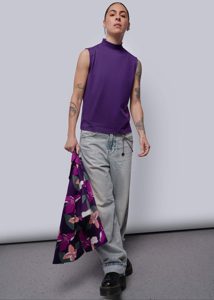 S=Person wearing a purple sleeveless top and gray pants, holding a floral shirt against a plain background.