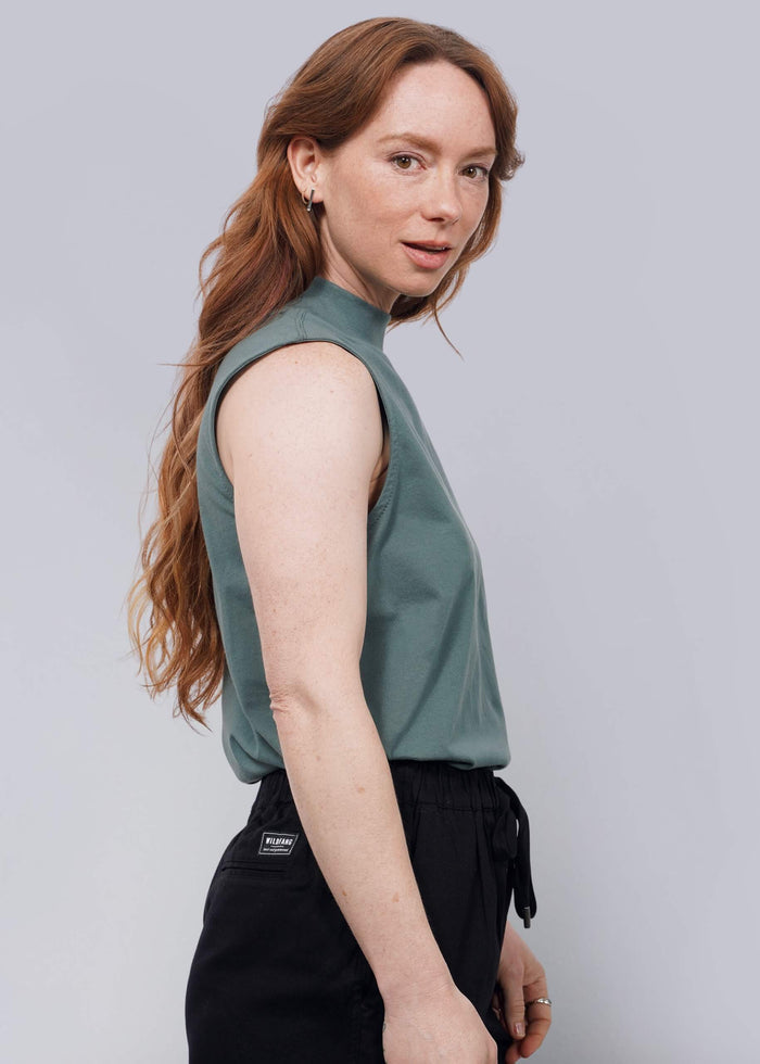 S=A woman with long, wavy reddish-brown hair stands sideways, facing the camera. She wears The Essential Mock Neck Tank in green and black drawstring pants against a plain white background.