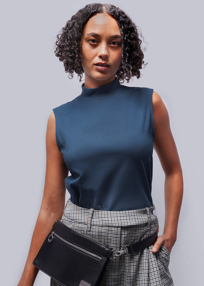 S=Woman wearing a dark blue sleeveless top and plaid pants, holding a black bag against a gray background
